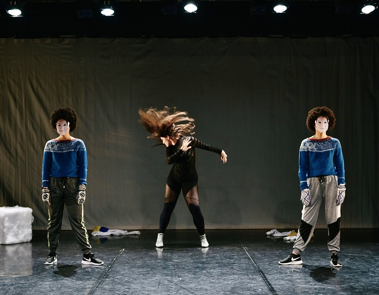 Two women with masks and Nordic sweaters stand like sentries on either side of a writhing dancer